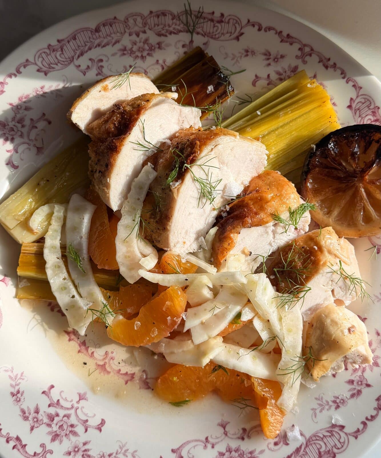 Sliced roasted chicken breast on a fennel and orange salad with braised leeks and a charred lemon half, served on a white floral-patterned plate.
