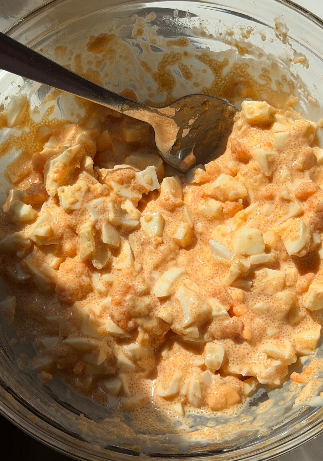 A glass bowl containing chopped hard-boiled eggs mixed with an orange-tinted creamy dressing, being stirred with a metal spoon.