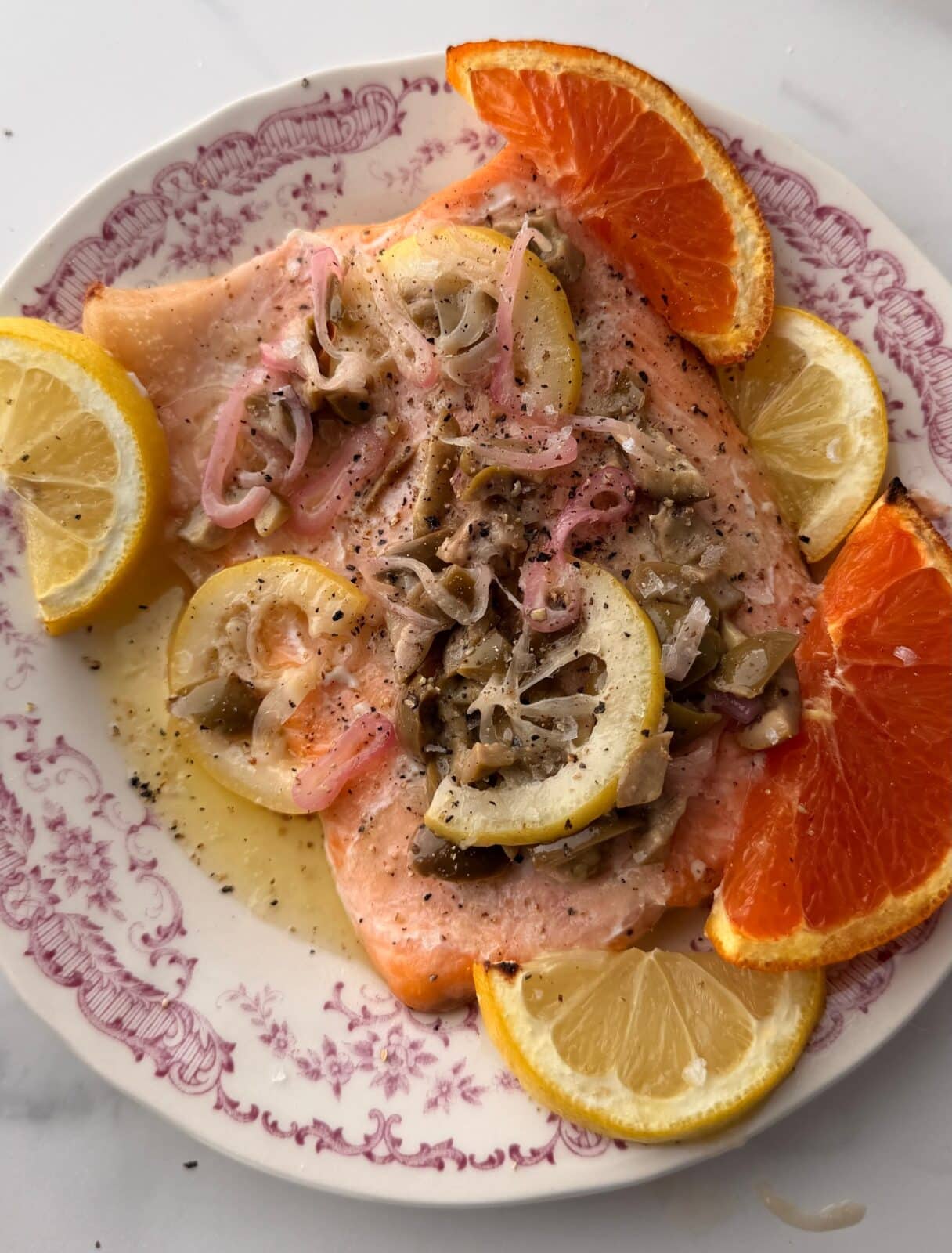 A plate with baked salmon topped with onions and mushrooms, garnished with lemon and orange slices on a white and pink patterned dish.