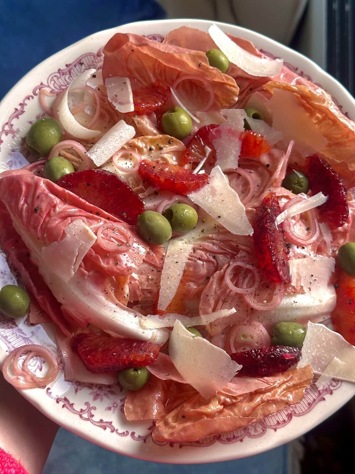 A plate of salad with red lettuce, green olives, sliced strawberries, shallots, and shaved cheese on a patterned dish.