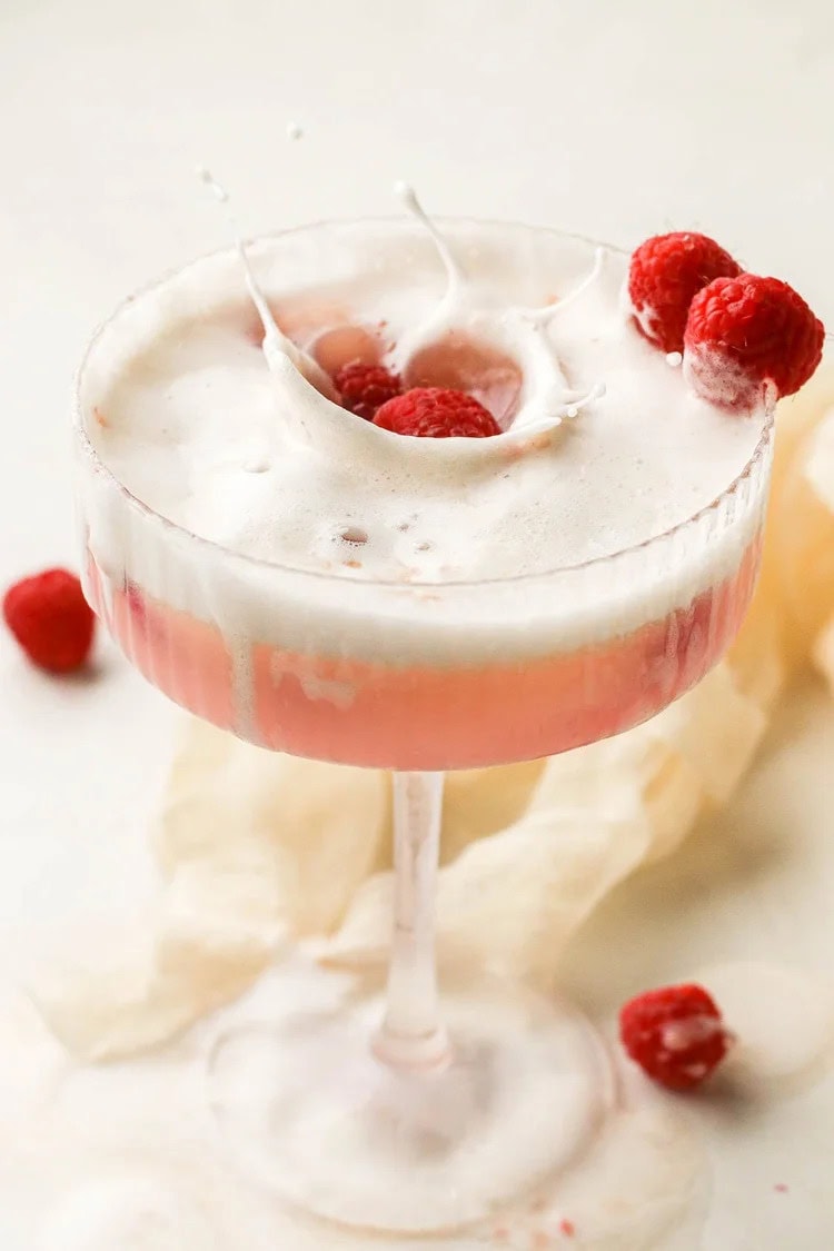 A pink cocktail with a frothy top in a coupe glass, garnished with raspberries. One raspberry is being dropped into the drink, creating a splash.