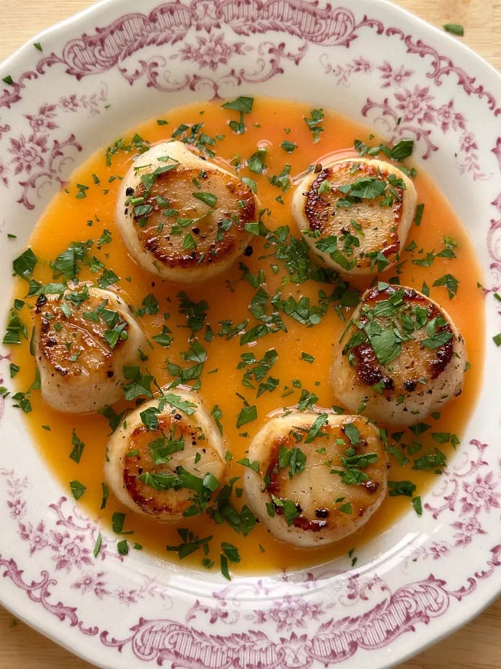 Seared scallops arranged in a patterned dish, topped with chopped herbs and surrounded by an orange-colored sauce.